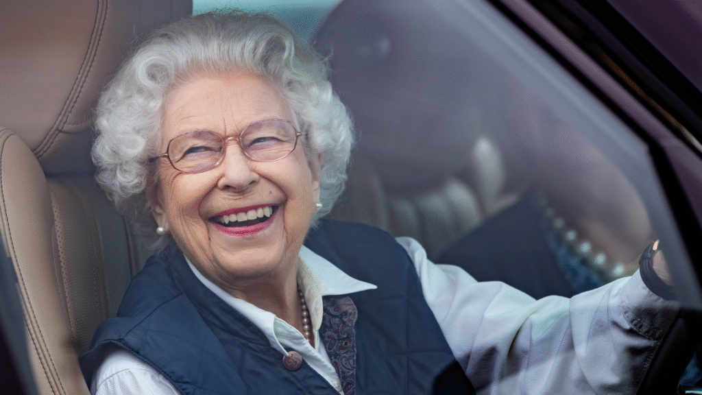 Queen Elizabeth wearing a blue vest and white shirt laughing while driving her car
