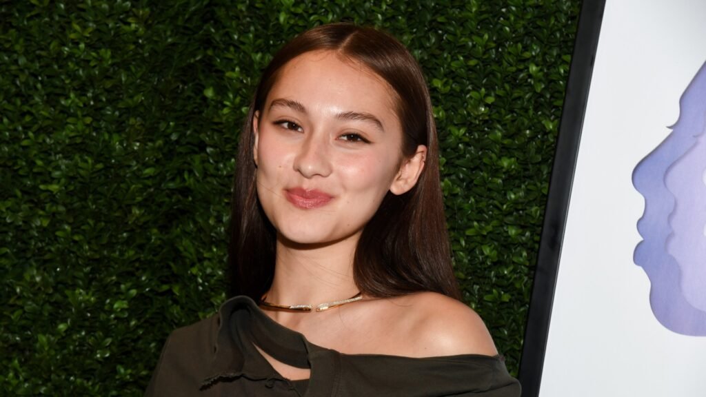 a woman with brown hair wearing an off-shoulder top posing in front of a grass wall