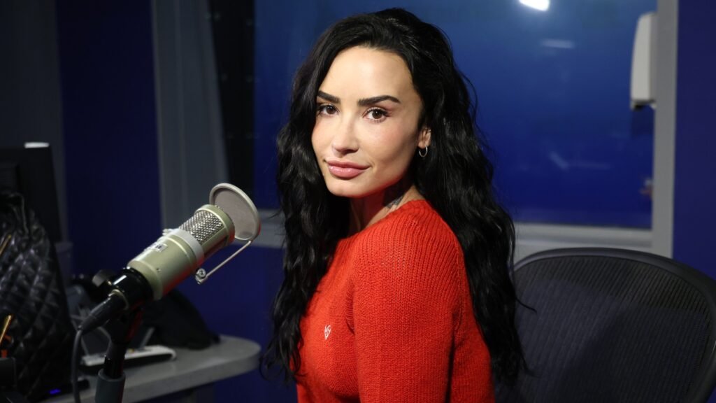 a white woman with dark hair wearing a bright red sweater and posing in front of a microphone in a studio