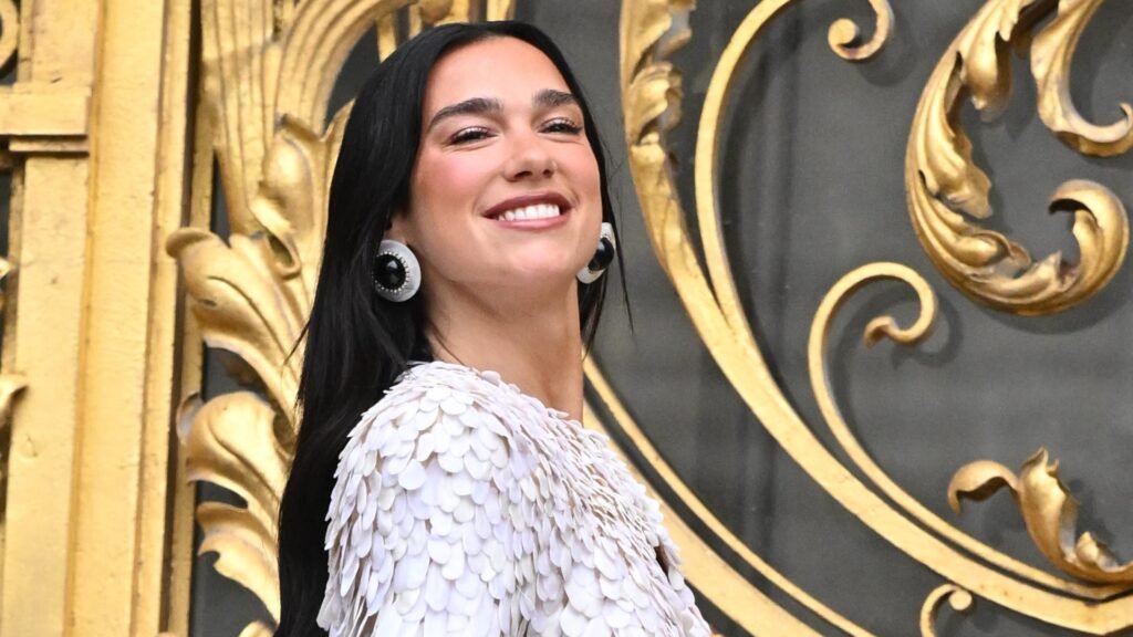 a woman with long, dark hair wearing a white, frilly top standing in front of gold doors