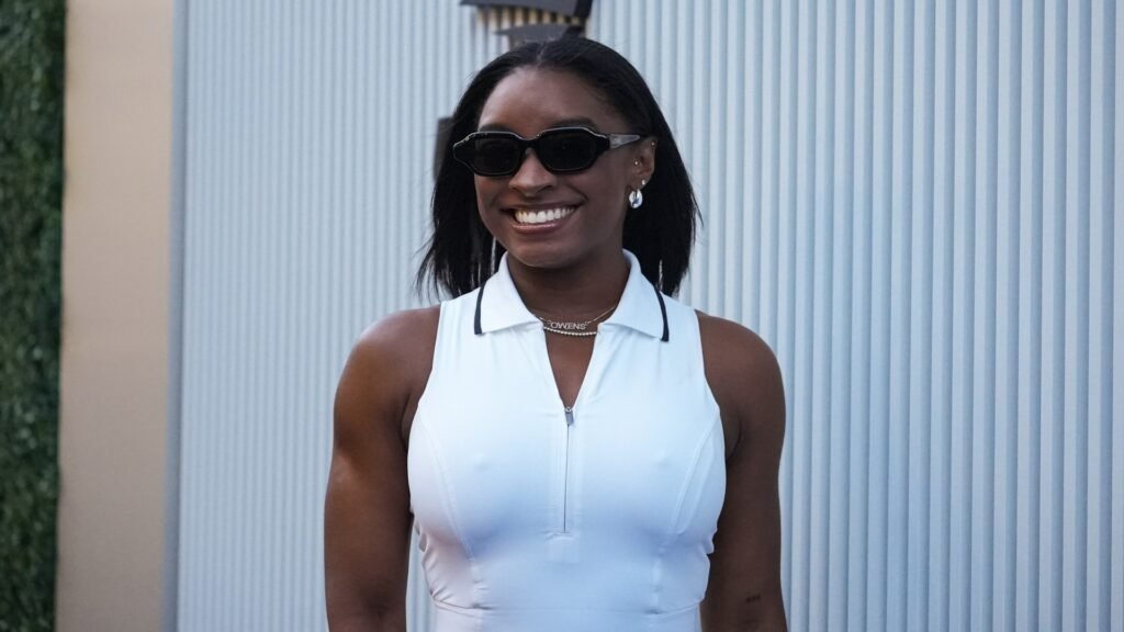 a black woman with dark hair wearing a sleeveless white shirt and black sunglasses smiling in front of a white background