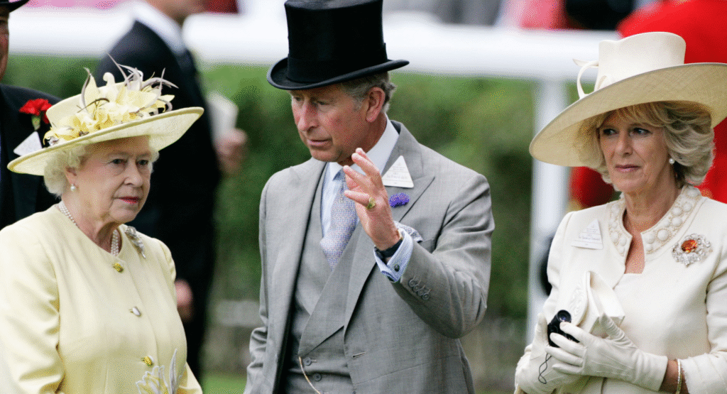 Queen Elizabeth II wears a pale yellow outfit while her son King Charles waves in front of her face, and Queen Camilla stands to the side ignored wearing white